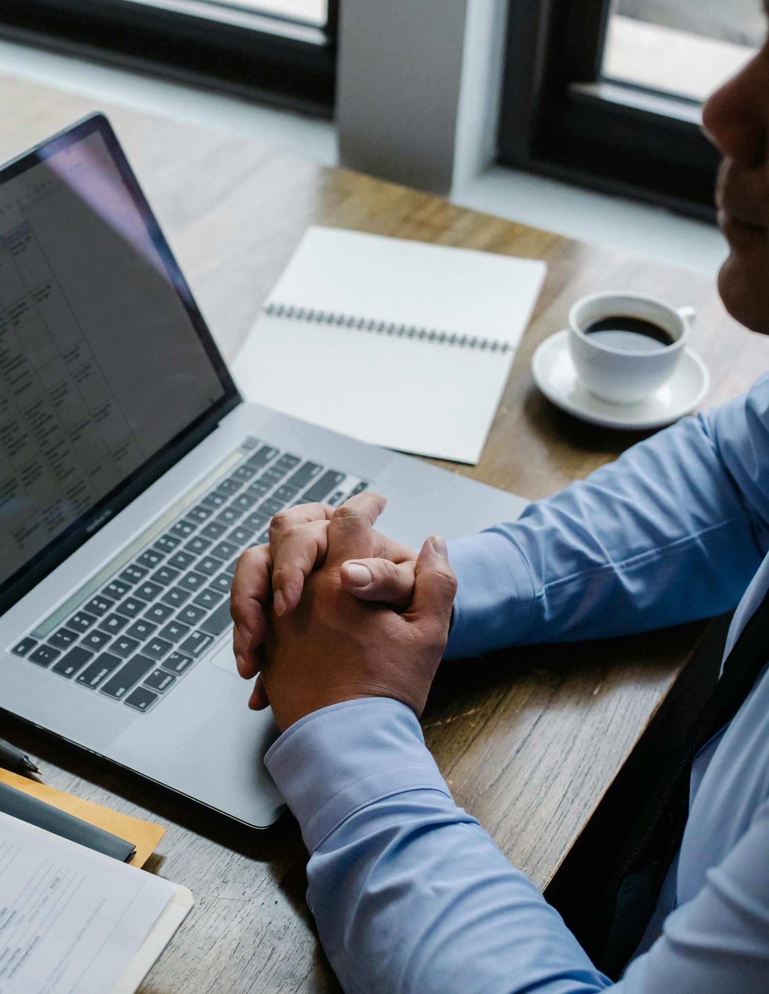 Man in blue shirt with hands clasped at laptop, coffee, and notebook